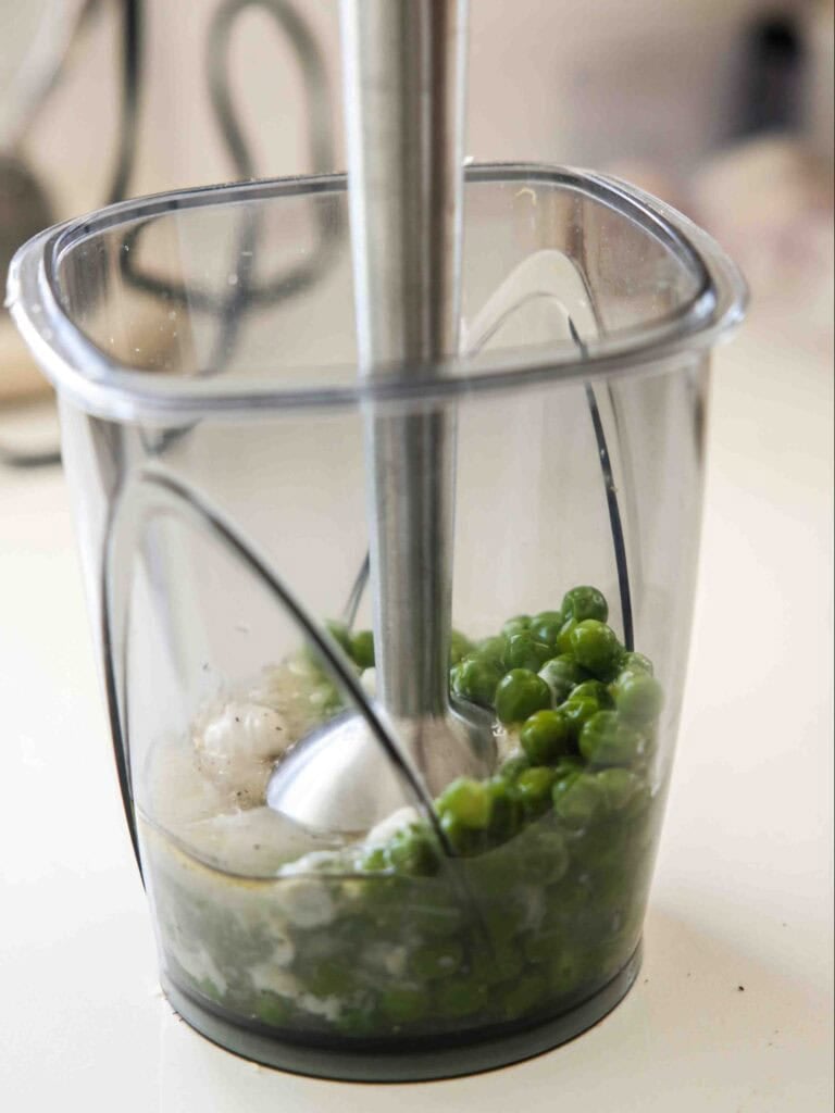 A handheld immersion blender is inside a clear plastic container, blending green peas and chopped onions on a kitchen counter.